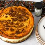 a top angle shot of a whole halloween cheesecake with pumpkin cheesecake top with chocolate swirls in it. In the scenery is Rodelle Gourmet Vanilla Beans, a stack of textured plates holding two vanilla beans on top, halloween fabric, and a piecutter.