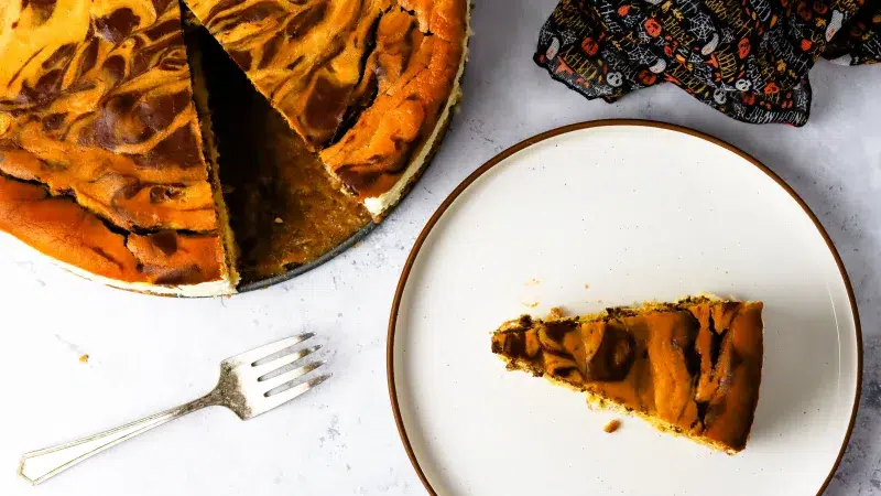 Top view of a halloween cheesecake. A slice has been taken out of it and lays on a plate. Surrounding the area is a fork and halloween fabric pattern.