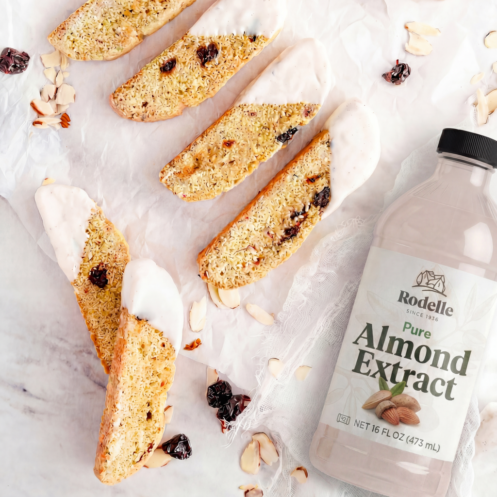 lifestyle recipe image of Almond and Cherry Biscotti Dipped in Vanilla Bean White Chocolate. some biscotties are neatly organized on parchment paper in the upper right. Rodelle 16oz almond extract bottle is in the bottom right. The background is a white and gray marbled pattern