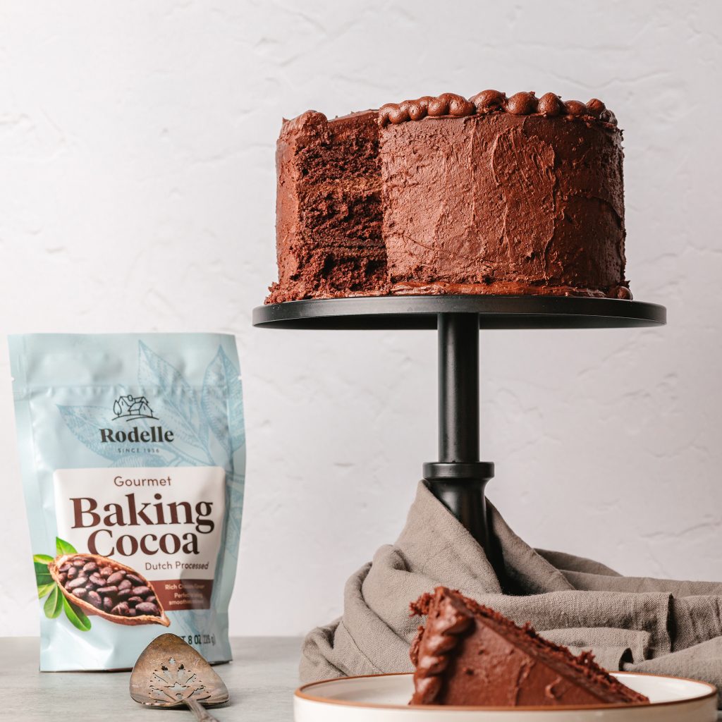 lifestyle shot of Devil’s Food Cake with Chocolate Espresso Frosting on a dark gray cake stand. In the background is Rodelle's 25oz gourmet baking cocoa bag. A slice of the cake is at the forefront. The background is a light gray