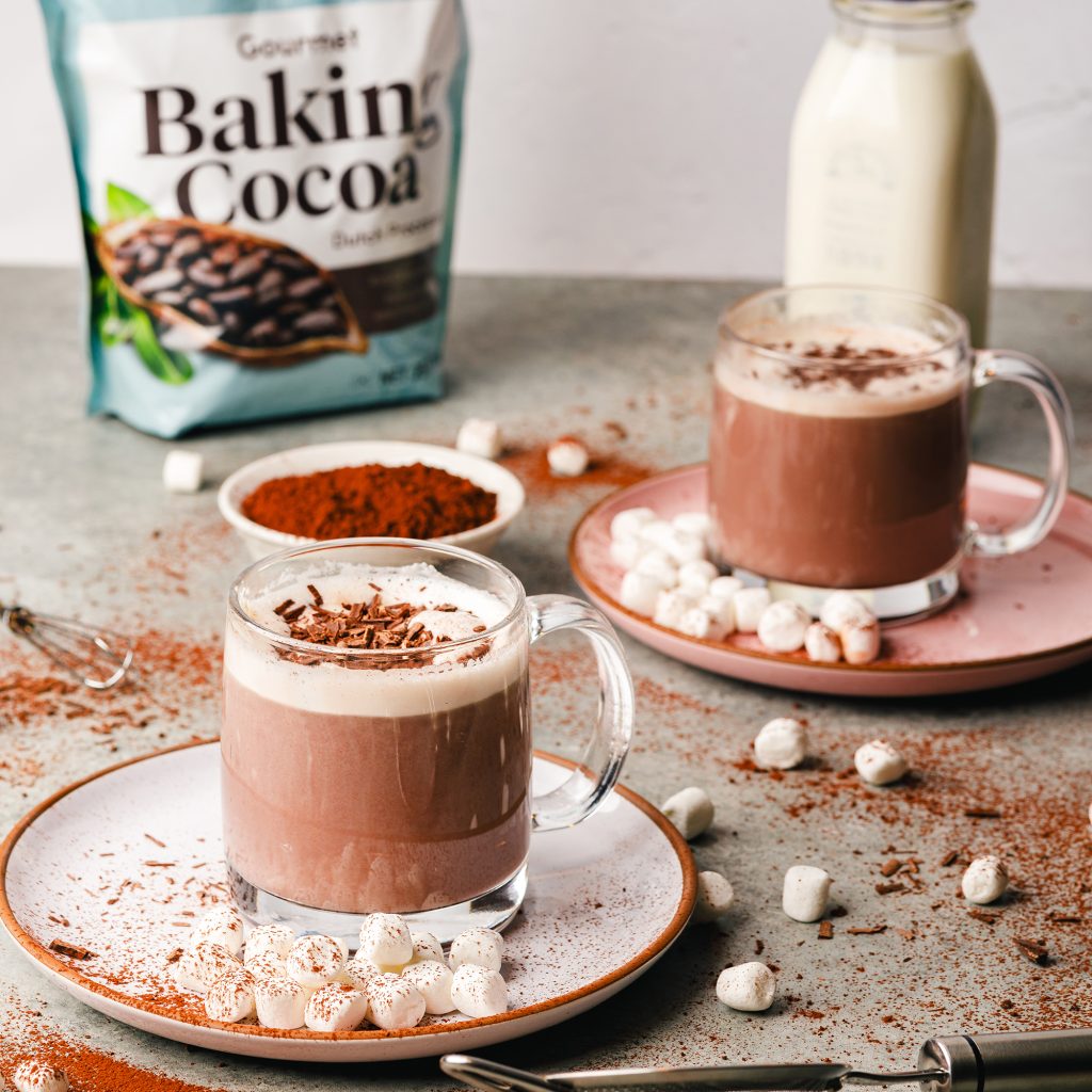 lifestyle image of a decadent hot cocoa in a clear glass mug. There are two mugs laying on earth toned plates. Each hot chocolate contains whipped cream and mini marshamallows which are also scattered around. shredded chocolate lays on top of whipped cream. In the background is Rodelle 25oz gourmet baking cocoa along with a small pint of cream