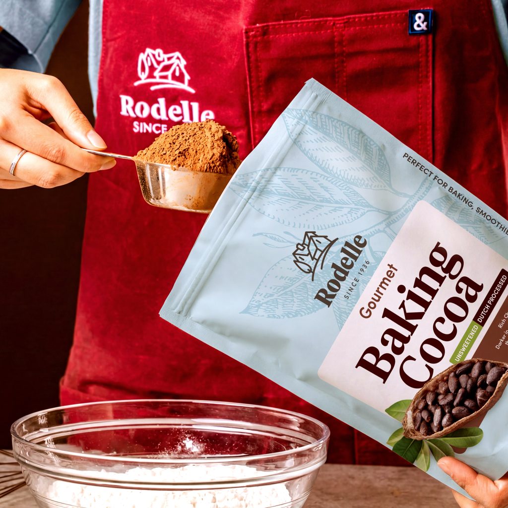 lifestyle image of a person with a metall measuring cup, scooping out baking cocoa from a Rodelle 25oz gourmet baking cocoa. The person is wearing a red apron with the Rodelle logo in white.