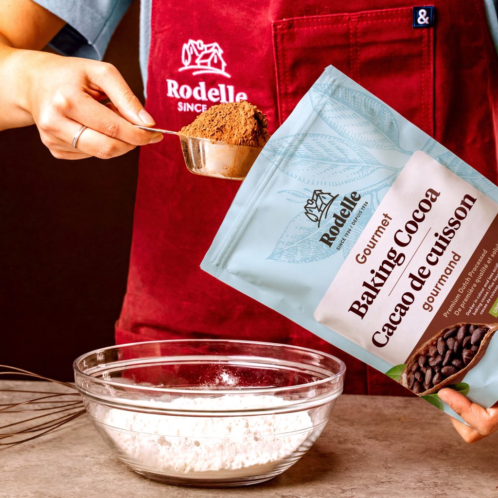 lifestyle image of a person with a metall measuring cup, scooping out baking cocoa from a Rodelle 25oz gourmet baking cocoa - canadian version. The person is wearing a red apron with the Rodelle logo in white.