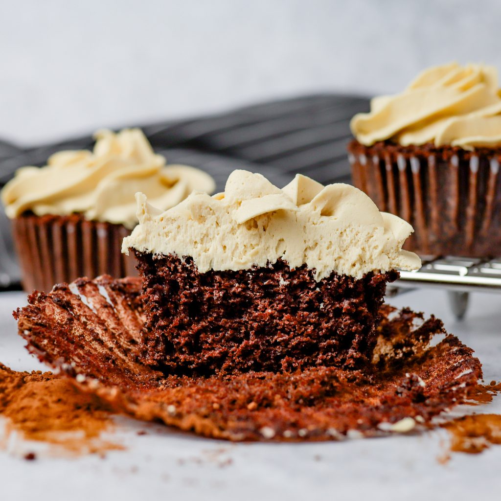 lifestyle image of Mocha Cupcakes with Espresso Buttercream Frosting. One cupcake is at the forefront and cut in half to see the texture of the cupcake. The background is blurry with two more cupcakes along with a dark gray towel. The table and most of the background is a light gray.