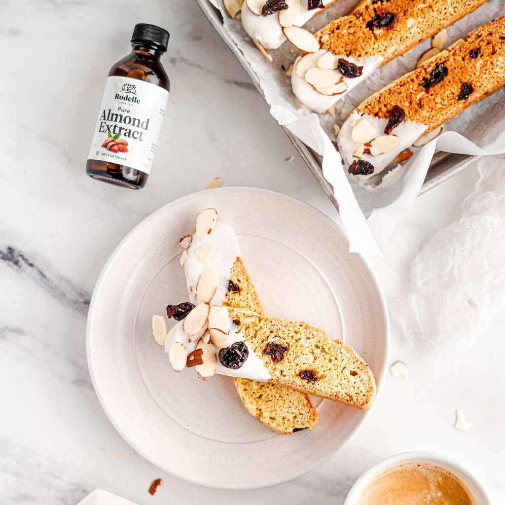 lifestyle recipe image of Almond and Cherry Biscotti Dipped in Vanilla Bean White Chocolate. some biscotties are in a pan in the upper right corner while one biscotti is on a small white plate. Rodelle 2oz almond extract bottle is in the upper right. The background is a white and gray marbled pattern