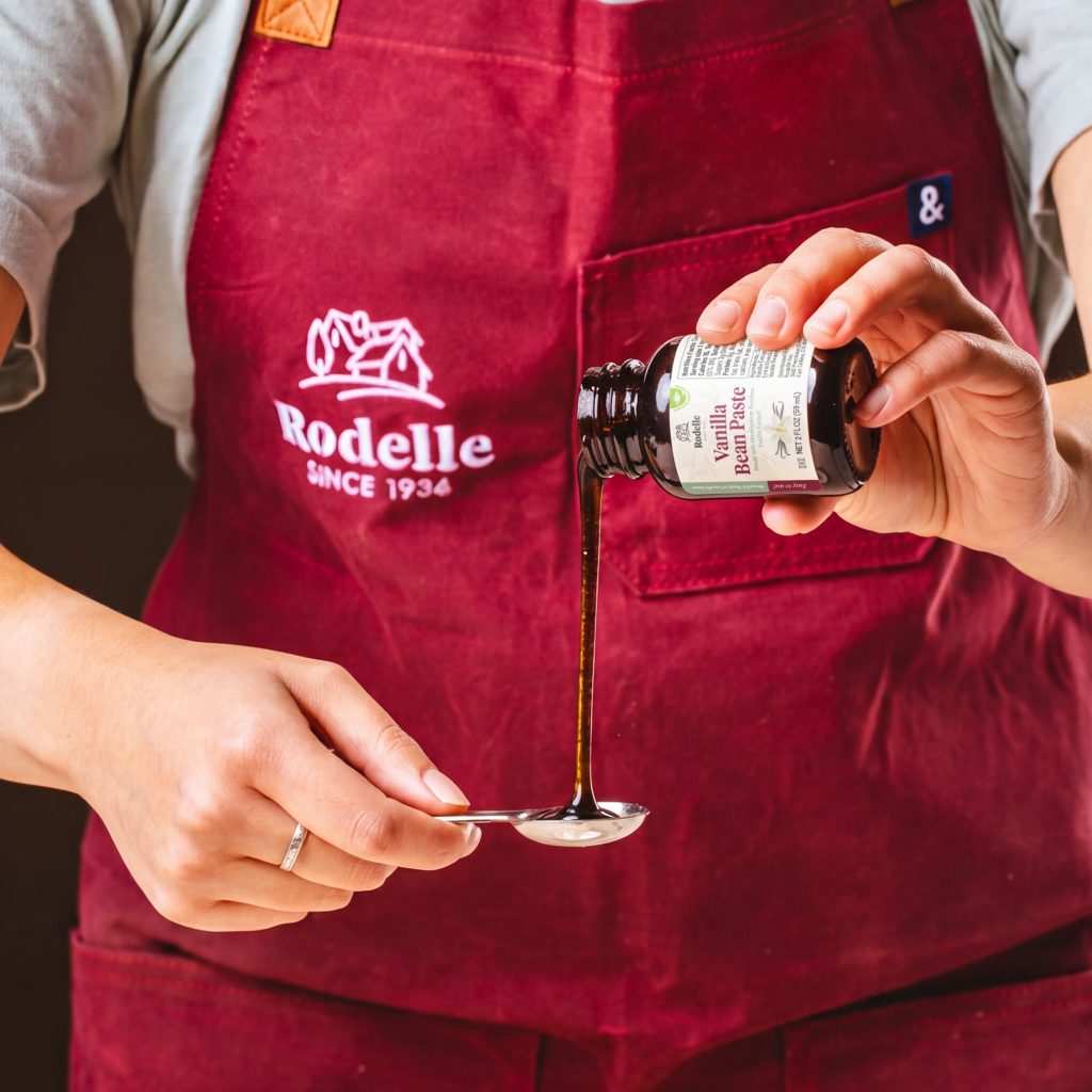 lifestyle image of a person pouring Rodelle's 2oz natural vanilla bean paste into a measuring spoon. the person is wearing a red apron with a white Rodelle logo