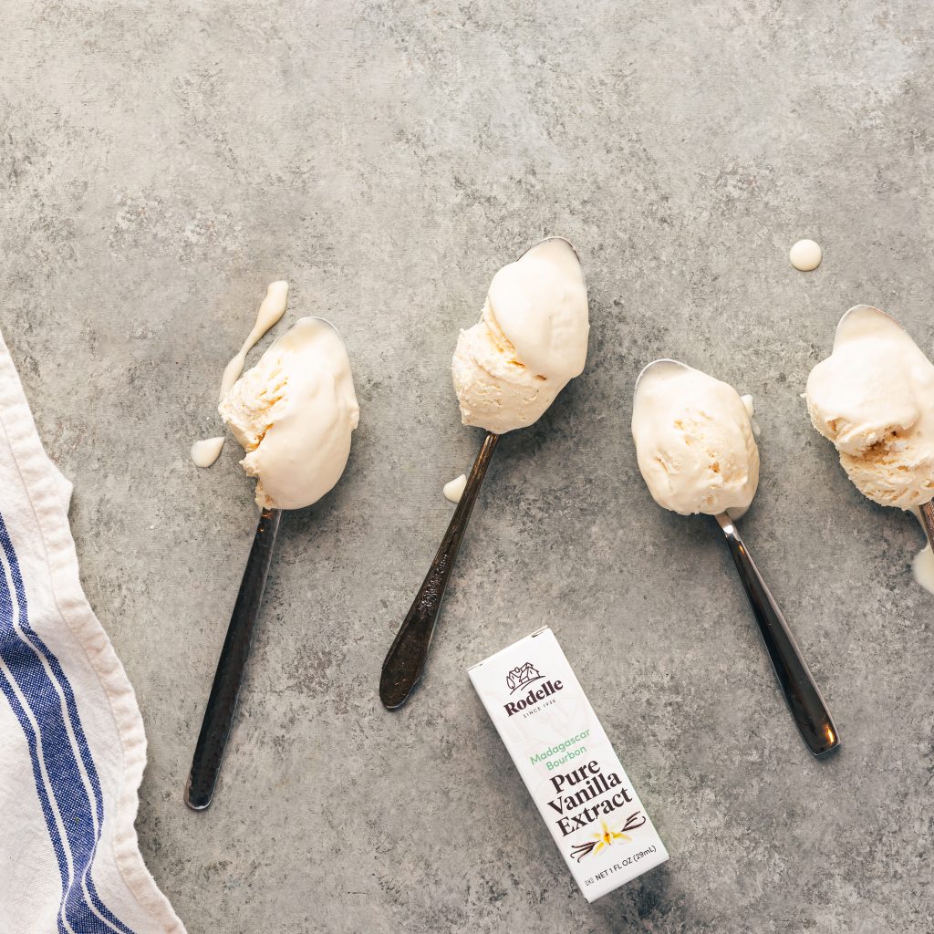 photograph of four silver spoons with scoops of vanilla ice cream. In the middle bottom area is a 1oz bottle of pure madagascar bourbon vanilla extract. To the left going off the edge is a white towel with one thick blue stripe as a accent. the table is dark gray.