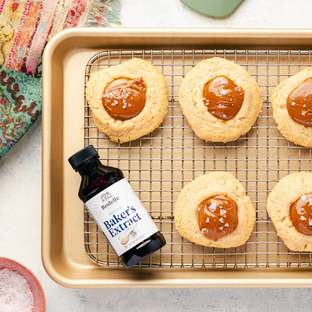 lifestyle recipe image of Salted Dolce De Leche Thumbprint Cookies. on a brass pan with a cooling rack are 5 thumbprint cookies and on the bottom left of the pan is Rodelle's 4oz Natural Baker's Extract. Surrounding the pan is a green towel and small bowl containing flaky salt - these go off the edge of the image