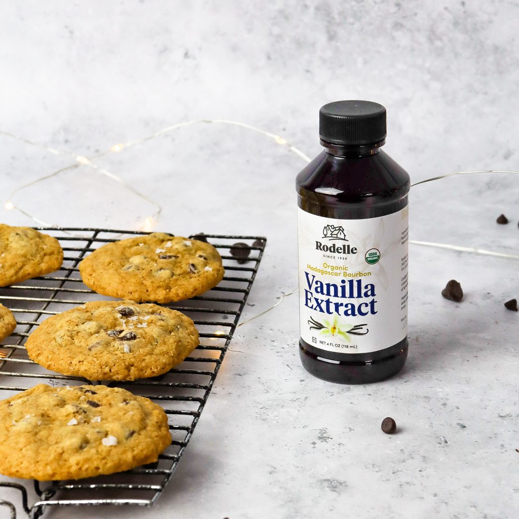 lifestlye image of Rodelle's 4oz Organic Pure Madagascar Bourbon vanilla extract next to a cooling rack containing homemade chocolate chip cookies. The background is a negural gray color and scattered around the image are chocolate chips.