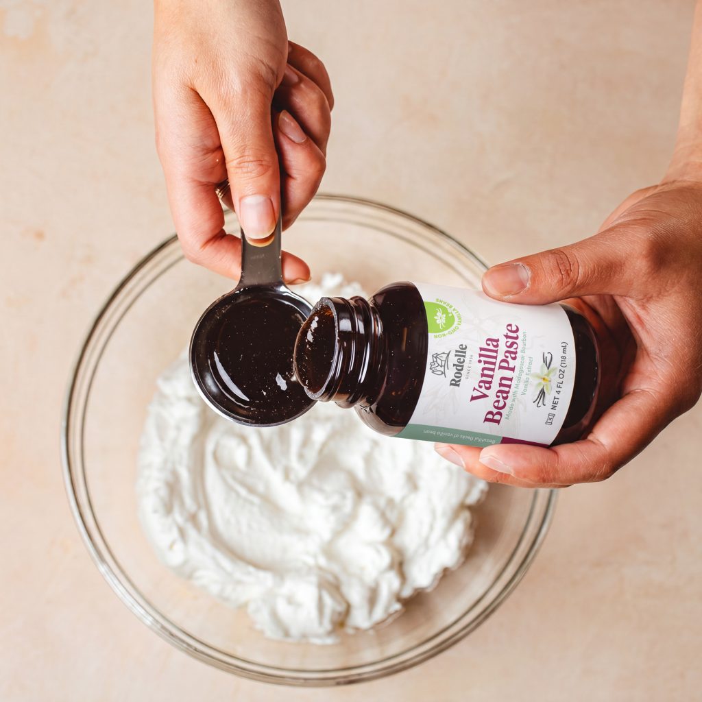 lifestyle image of hands pouring 4oz natural vanilla bean paste into a tablespoon. Below the vanilla paste and hands is a clear bowl is whipped cream. the table is a muted peach color