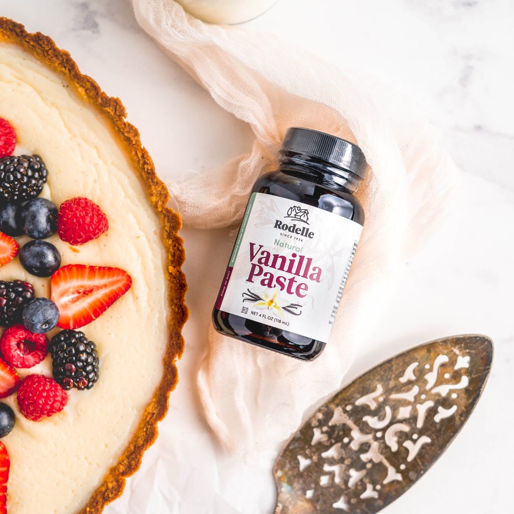 recipe image of rodelle 4oz natural vanilla paste laying on a light pink towel. To the left is a vanilla bean berry custard tart and to the bottom right is rustic pie cutter. in upper part of the image is a glass of milk. the table is a white marble pattern