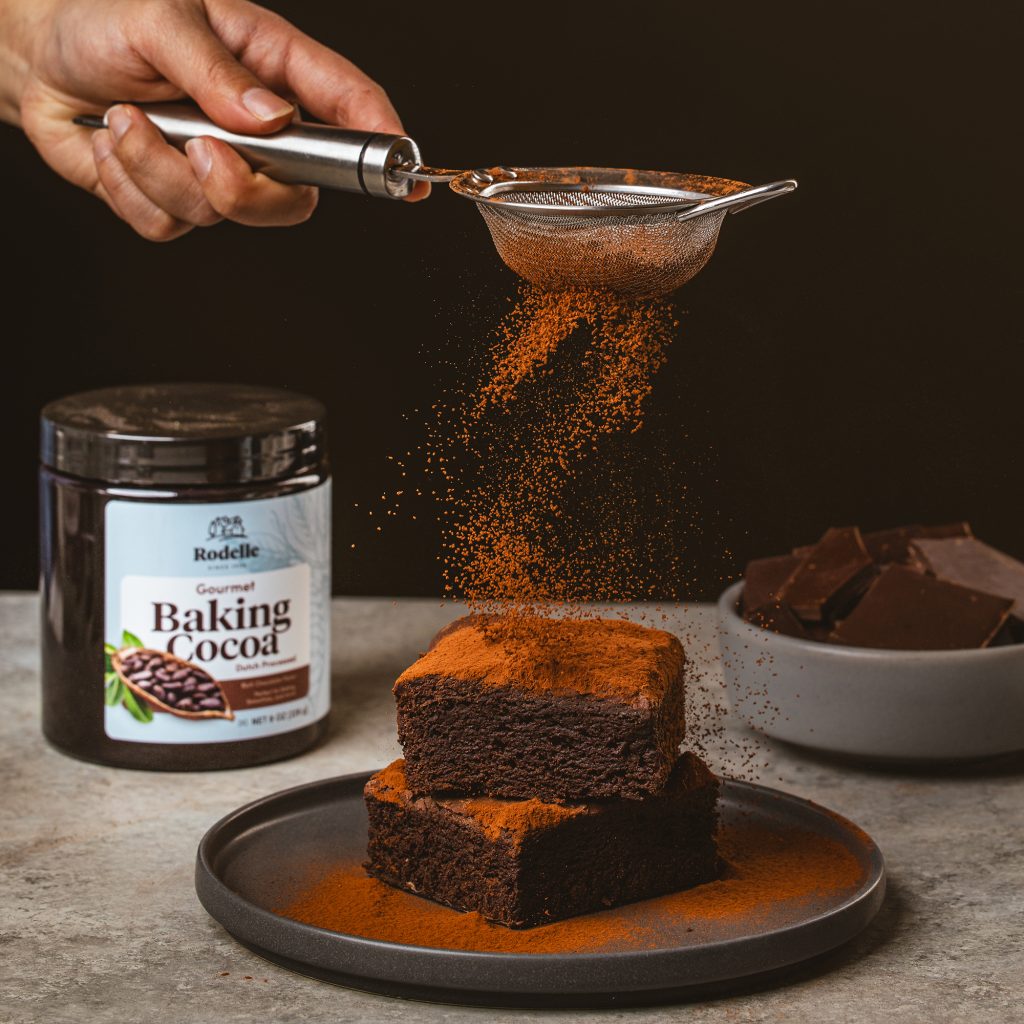 lifestyle shot of baking cocoa being dusted onto brownies on a dark gray plate. the background is black and to the left side is Rodelle's 8oz gourmet baking cocoa in a canister
