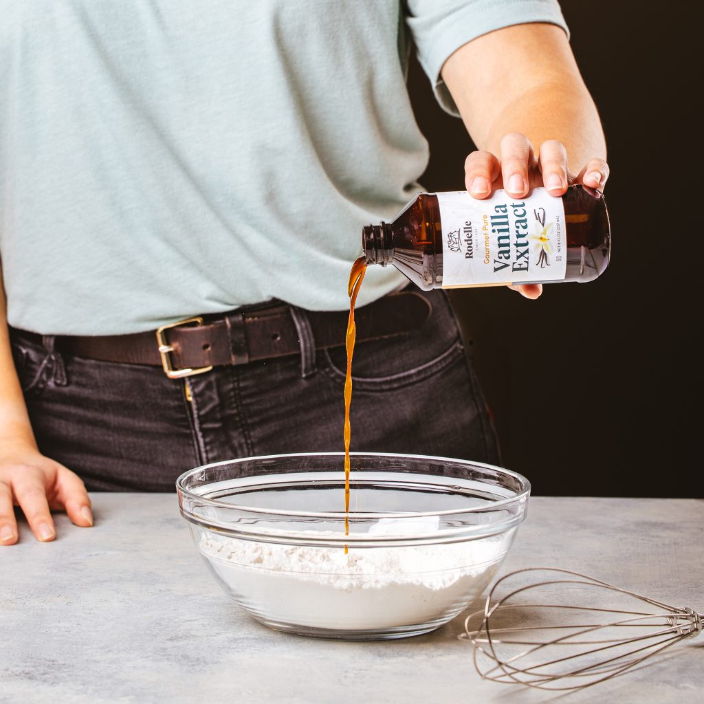 lifestlye image of someone in a muted sage green shirt pouring vanilla from a Rodelle 8oz gourmet pure vanilla extract bottle into a clear bowl with flour.