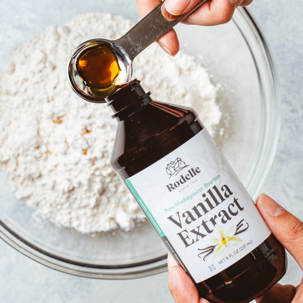lifestyle image of someone pouring Rodelle 8oz madagascar bourbon pure vanilla extract into a measuring spoon. underneath the vanilla extract being poured is a clear mixng bowl with flour. the background is a neutral blue and light gray.