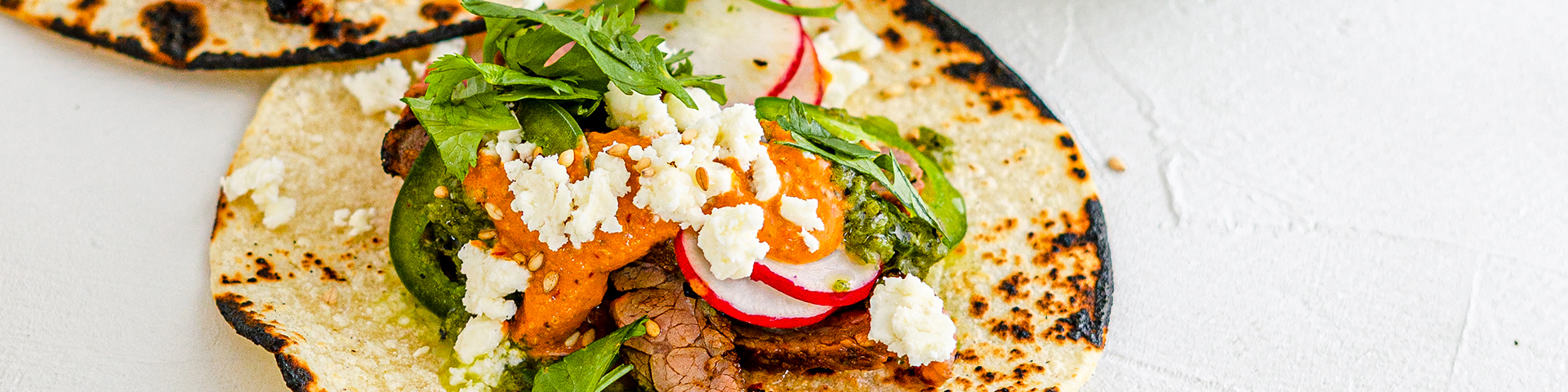 Close up of two prime tacos containing steak pieces, homemade salsa verde, toasted pepita salsa rojo, and topped with diced onion and cilantro. Tacos lay on a light beige plate with a beige napkin surrounding them. In the background is also Rodelle Prime Rib Seasoning and Rodelle Toasted Sesame Seeds.
