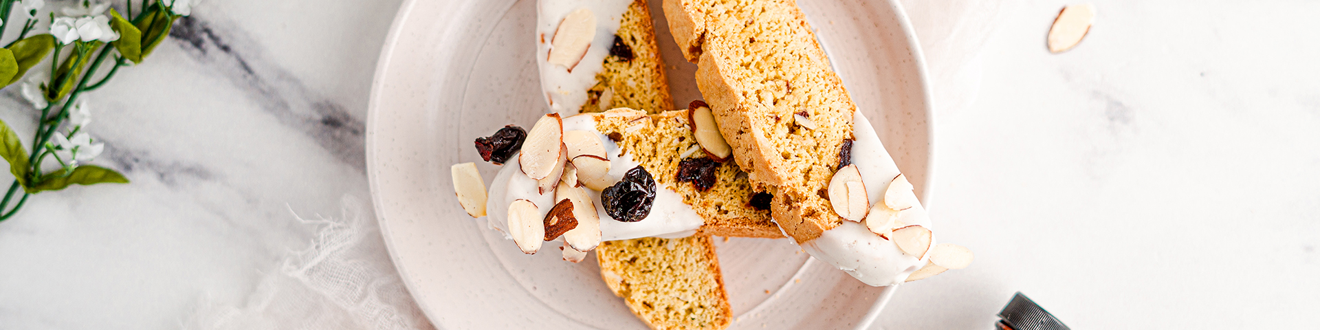 top down view of three Almond and Cherry Biscotti Dipped in Vanilla bean White Chocolate. they lay crossed over on a small whtie plate. The table is white and in the upper left is some green decor.