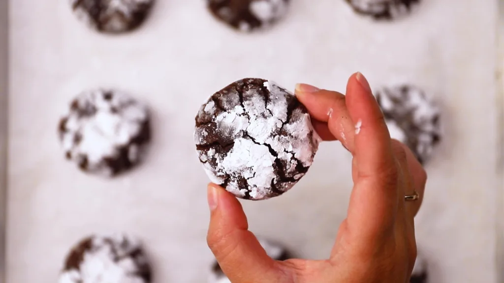 top image of a hand holding a chocolate crinkle cookie (with powdered sugar). Below the hand are additional cookies on parchment paper