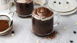 Dark and rich decadent hot cocoa served in a clear glass mug topped with swirl of whipped cream and dusted with baking cocoa. Surrounding the mug is a small sifter with baking cocoa, white and dark chocolate chips, white plates, and another mug of hot cocoa in the back.