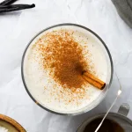 Top down shot of double vanilla eggnog with a dusting of cinnamon on top and a cinnamon stick sticking out. Surrounding the glass are vanilla beans, granulated white sugar in a brown ceramic bowl, and a sifter holding cinnamon.