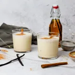 Side profile of two glasses of double vanilla eggnog laying on crinkled parchment paper. Surrounding the glasses is a homemade vanilla infused alcohol bottle, a gray towel, vanilla beans, cinnamon stick, a sifter, and a brown ceramic bowl holding granulated white sugar.
