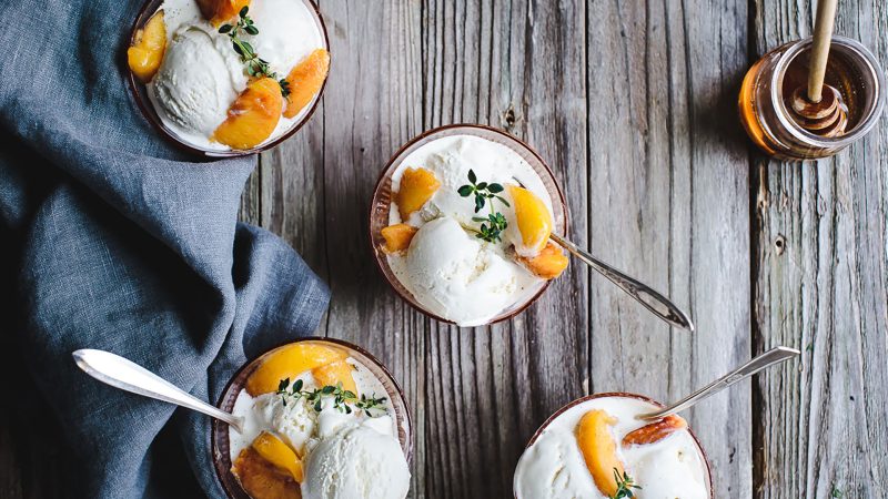 top shot of four small bowls containing honey lemon thyme ricotta ice cream with vanilla peaches. All bowls contain silver spoons and the counter the ice cream is on is a pale cool wooden color. To the left is a blue tow and upper right a small jar of honey with honey want.