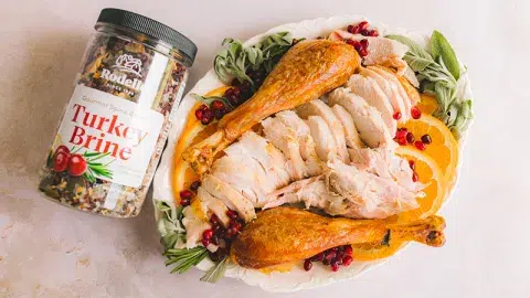 Top view of a roasted turkey cut into slices with two turkey legs on the size, orange slices, and pomegranate seeds. Laying next to the turkey plater is Rodelle's Turkey Brine.