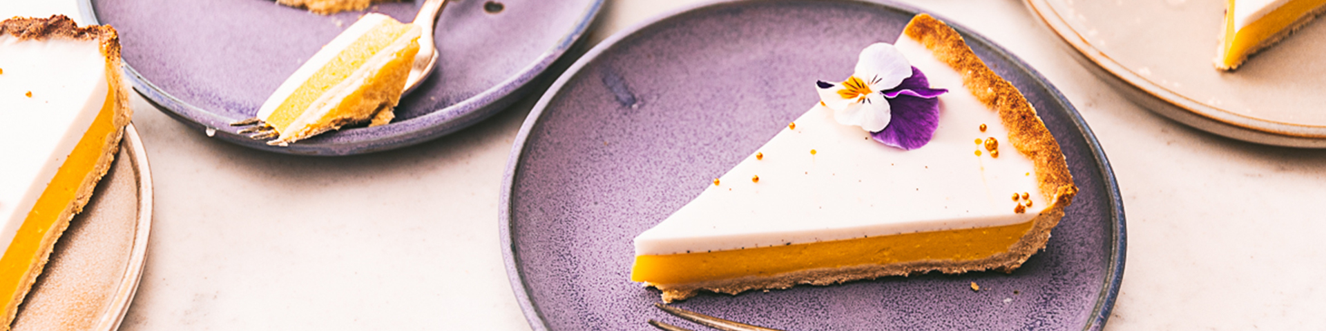 Mango tart with vanilla coconut custard sliced on neutral background with edible flours on purple plates