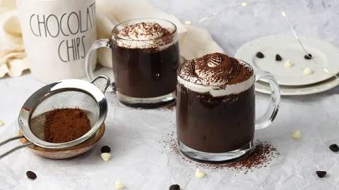 Dark and rich decadent hot cocoa served in a clear glass mug topped with swirl of whipped cream and dusted with baking cocoa. Surrounding the mug is a small sifter with baking cocoa, white and dark chocolate chips, white plates, and another mug of hot cocoa in the back.