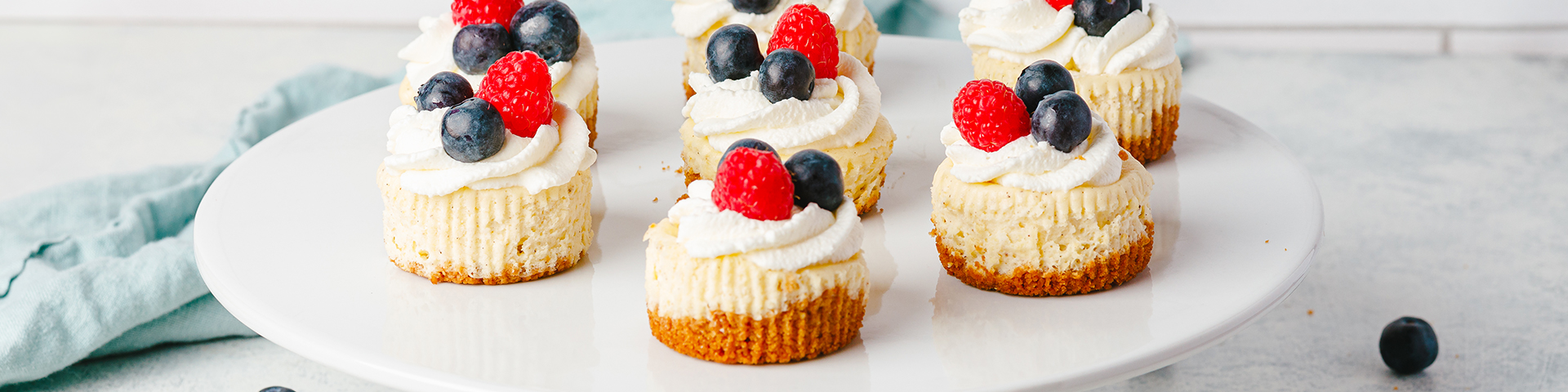 side profile of multiple mini vanilla bean cheesecakes on top of a white cake stand. In the background is white tile, a light turquoise blue towel, and in the foreground is a group of large strawberries. Topping the mini cheesecakes is a swirl of whipped cream, raspberries and blueberries.