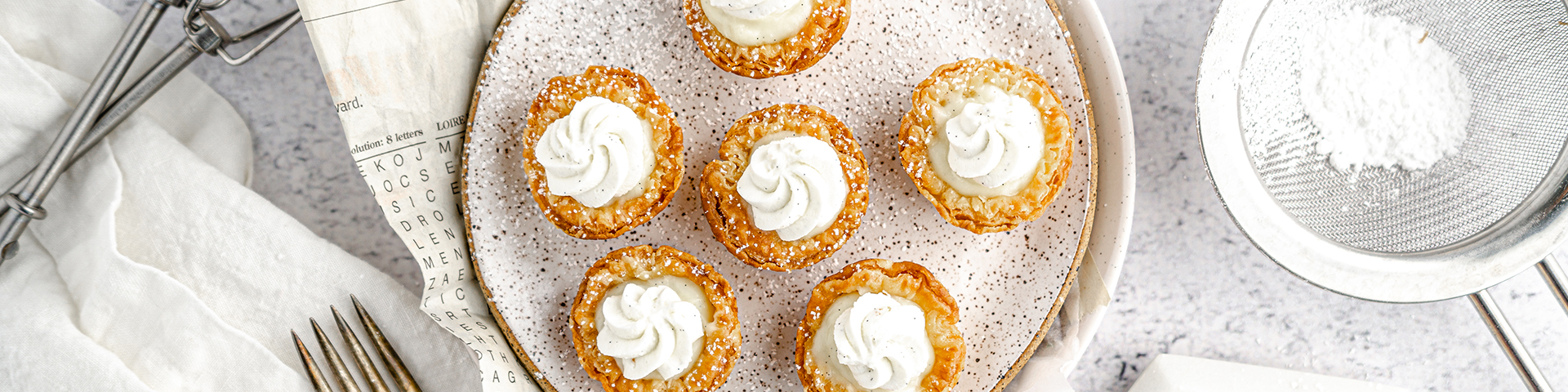 vanilla cream pies on neutral background shown from overhead with plates, serving utensils and pwodered sugar