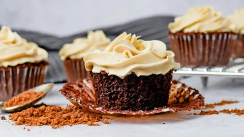 Mocha cupcake with espresso buttercream piped on top of them. One of the cupcakes is unwrapped showing. Surrounding the unwrapped cupcake is baking cocoa scattered and laying on a spoon. Int eh background are more cupcakes and a gray striped fabric..