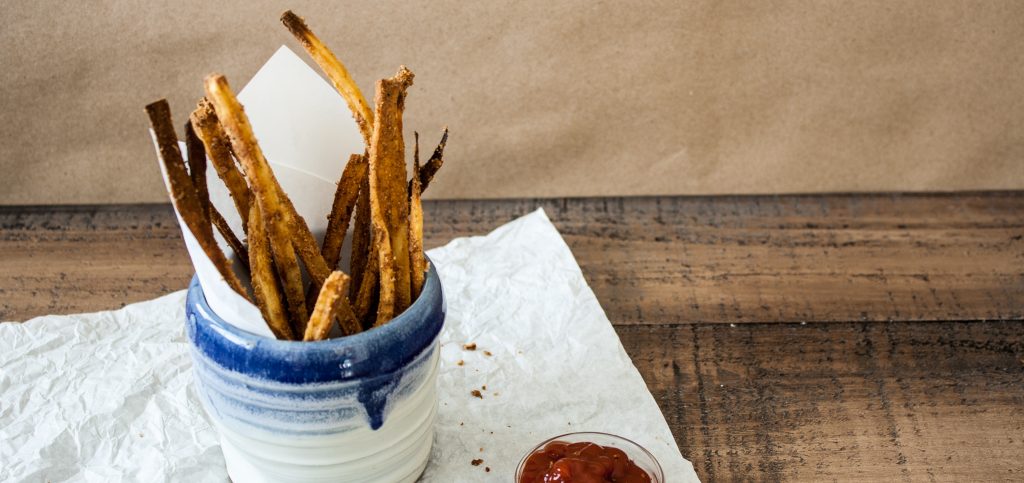 lifestyle image of parsnip fries in a cute pottery white and blue cup. The ceramic cup lays on parchment paper. the background is wooden and tan.