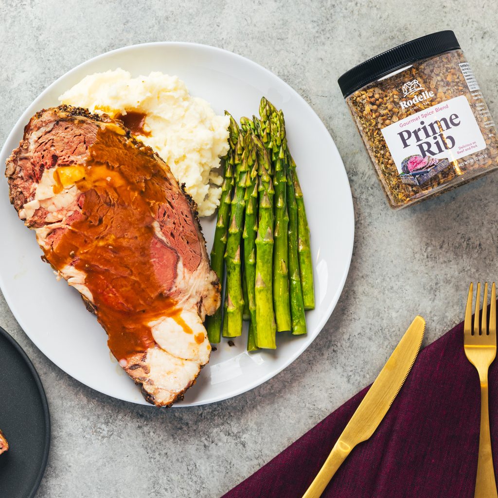 lifestyle image of a platter with a slice of prime rib, cooked asparagus, and mashed potatoes. surrounding the plate is a dark purple towel with brass colored utencils. In the upper right is Rodelle's 15oz prime rib seasoning on it's side