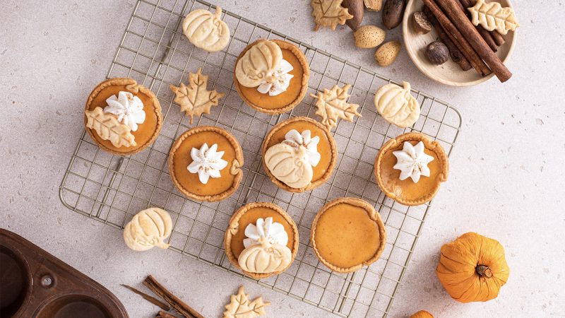 top view of pumpkin spice tartlets laying on a cooling rack. Each tartlet has whipped cream and a pumpkin shortbread cookie. Surrounding the tartlets are fall themed items like cinnamon sticks, acorns, more shortbread cookies, and a rustic pan.