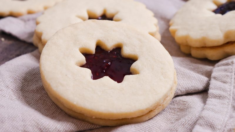 close up shot of a circular jam filled shortbread cookies on a light purple brown kitchen towel. The cooke on top of the jammed filled cookies is a star/flower pattern and a lighter strawberry jam color stick out of it.