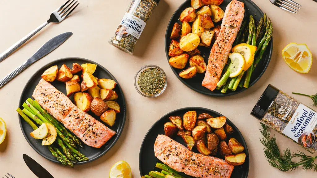 Overhead shot of salmon and veggie seafood sheet pan recipe. Instead of a sheet pan, the salmon, small potatoes, and asparagus with slivers of lemon have been served onto three charcoal matte ceramic plates. The background is a light orange salmon color. Rodelle's retail sized seafood seasoning and larger size lay on their sides with label facing upwards. Slices of lemon, pinch bowl with seafood seasoning, dill, and silver cutlery surround the plates.