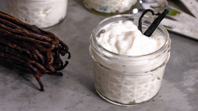 angled photo of a small mason jar with vanilla bean pudding. You can see the flecks of vanilla seeds in the pudding along with two vanilla beans sticking out of the jar. To the left of the jar is a bundle of vanilla beans. Int he background are additional mason jars filled with pudding and measuring spoons. The counter is a cookie sheet/metal texture