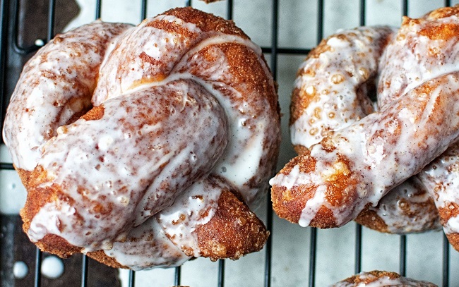 close up shot of vanilla bean glazed soft pretzel donuts on a cooling rack. below them is a cooler toned tray/background.
