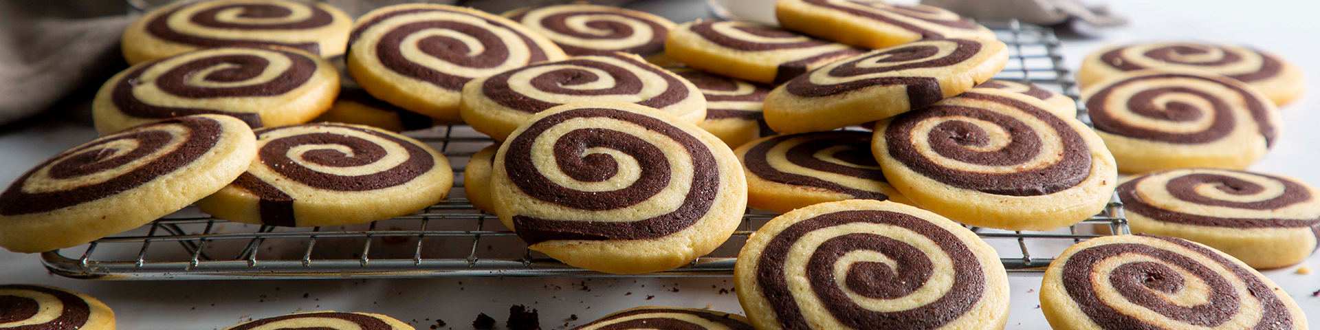 Vanilla-Chocolate Pinwheel Cookies shown on a cooling tray with cups of milk and cloth in background