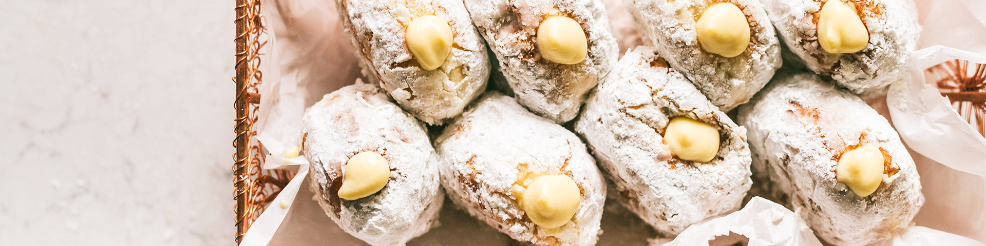 Fluffy donuts that are filled with a vanilla custard and doused in powdered sugar shown from an overhead angle in parchment paper and serving basket