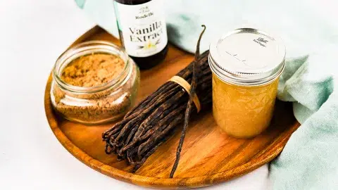 Large wooden plater containg a mason jar with vanilla infused body scrub, vanilla bean bundle, jar of sugar, and Rodelle Pure Vanilla Extract. Light blue towel lays on the side