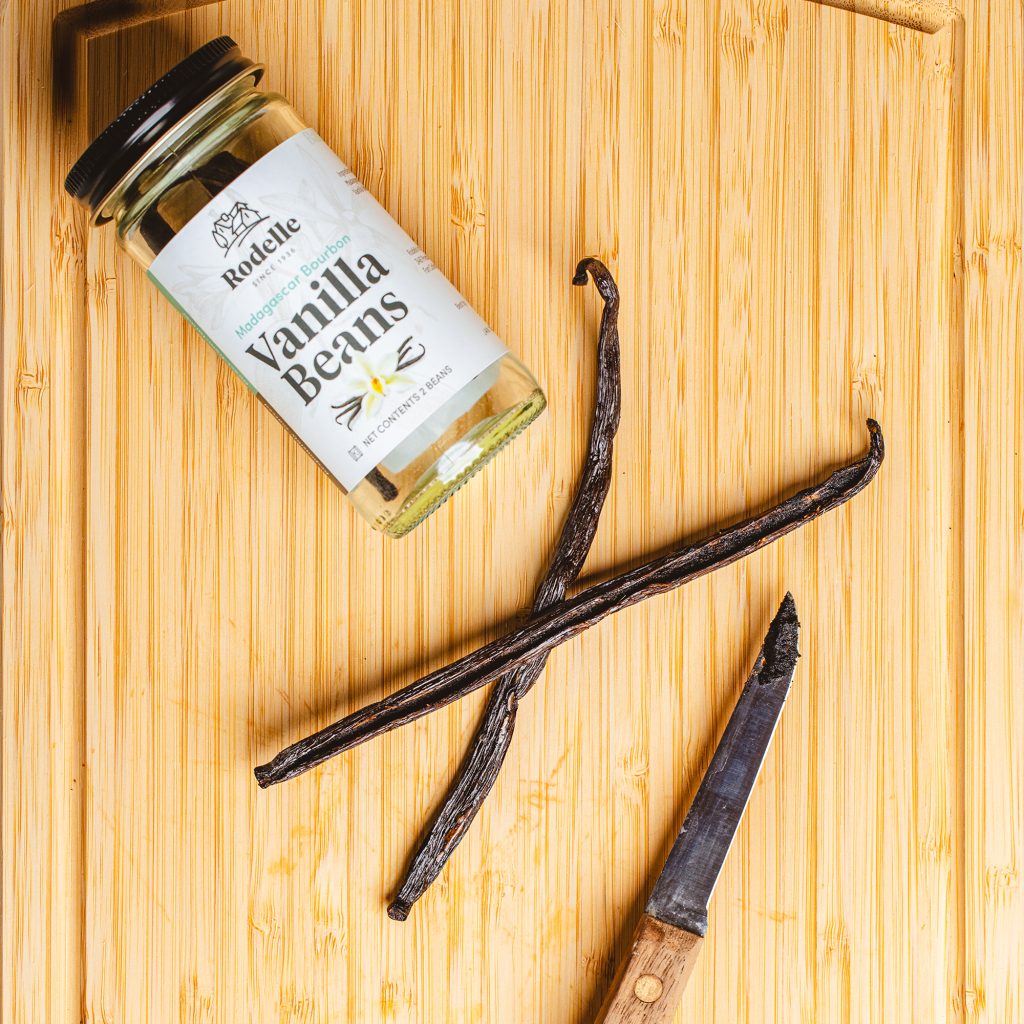 lifestyle image of rodelle 2ct madagascar bourbon vanilla beans on it's side on a cutting board. the jar is in the upper left. in the center is a sliced vanilla bean and you can see the vanilla seeds in it. to the right of the vanilla beans is a small knife