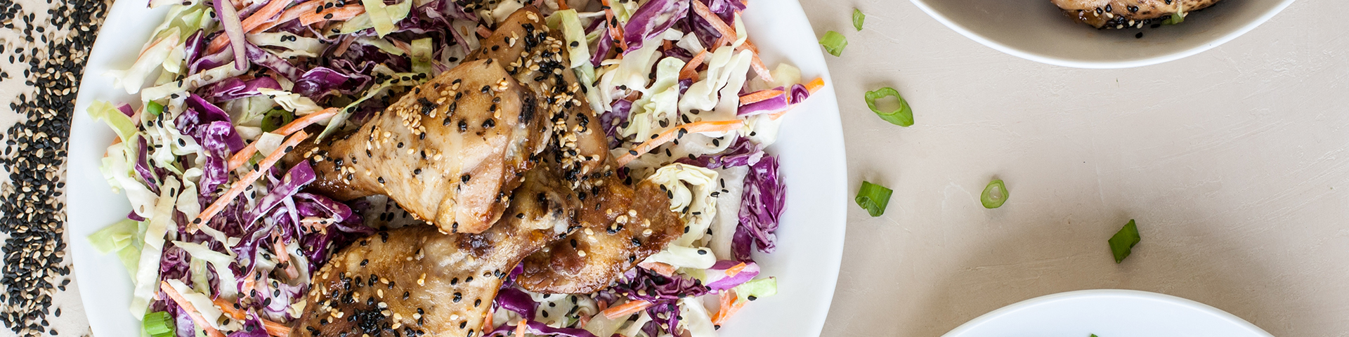 bowls of sesame chicken drumsticks shown from overhead angle with cole slaw and a scattering of black sesame seeds, toasted sesame seeds and chopped green onions