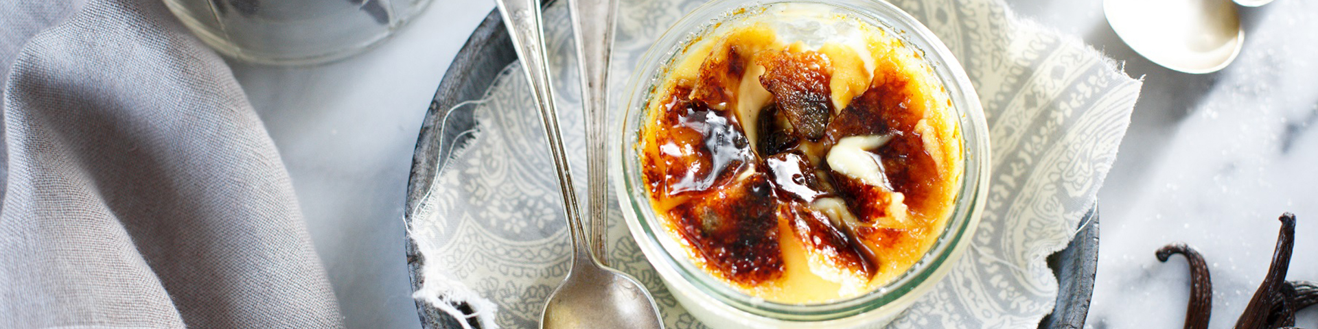 bowl of crème brulee shown from overhead with two silver spoons, the crakly top broken open and small bundle of vanilla beans