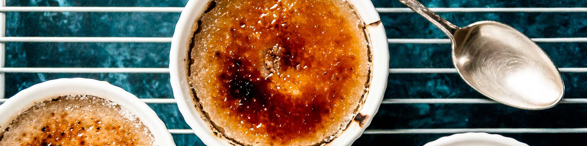 bowl of crème brulee shown from overhead with two silver spoons, the crakly top broken open and small bundle of vanilla beans
