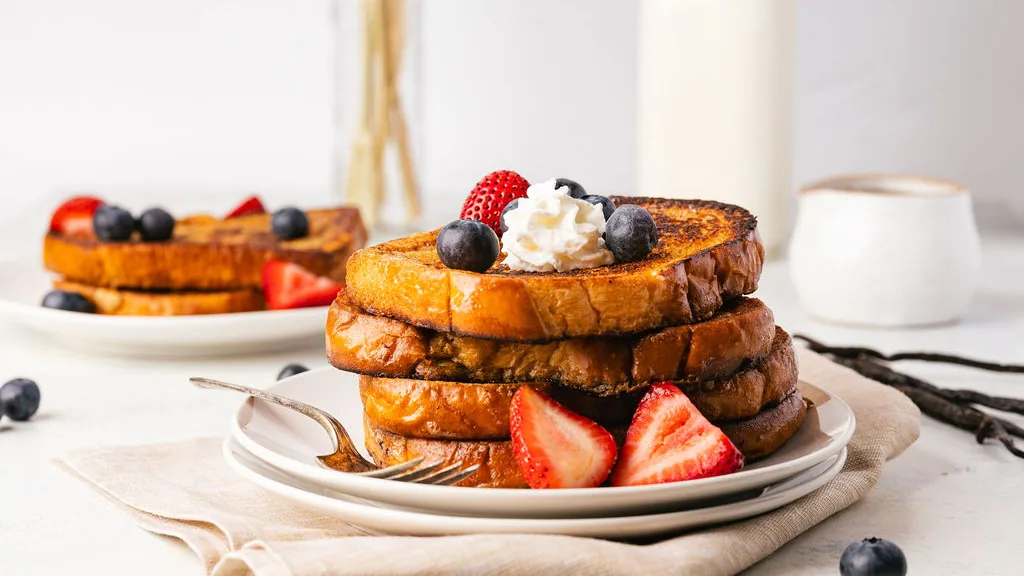 close up side shot of a large stack of bourbon vanilla french toast on a white plate stacked on another plate. Underneath the plate is a light tan cloth napkin. On the stack of french toast are blueberries, strawberries, and a bit of whipped cream. In the background is another stack of french toast, vanilla beans, pint of milk, yellow grass decor, and a small speckled jar for pouring containing maple syrup.