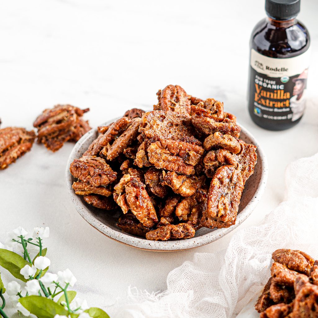lifestlye image of Vanilla Spice Caramelized Pecans in a small white bowl. in the bottom are smalle white flowers with small greeen leafes. in the upper right is a bottle of Rodelle 2oz organic fairtrade pure vanilla extrac