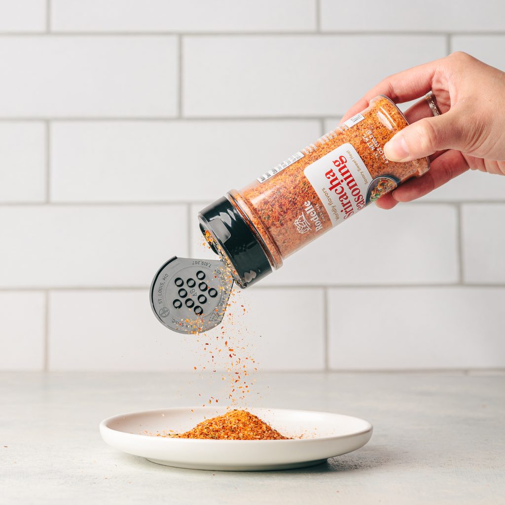 lifestyle image of a hand pouring Rodelle 4.5 sriracha seasoning on an white plate. the background is white tile