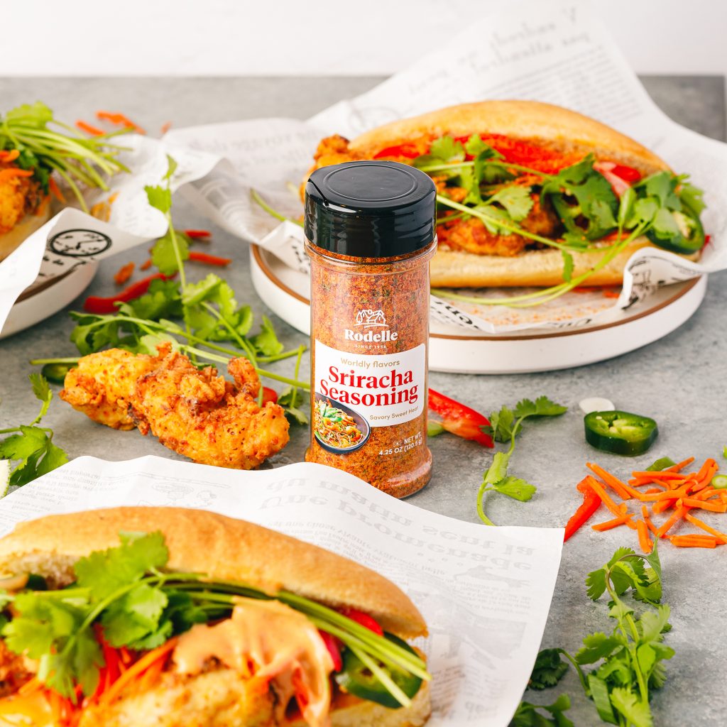 recipe image of sriracha fried chicken bahn mi sandwiches on earth toned speckled plates with brown edges. on the sandiwhces is fried chicken, pickled carrots, sriracha mayo, cilantro, and jalapenos. In the middle of the image is Rodelle 4.5oz sriracha in a round bottle with black cap.