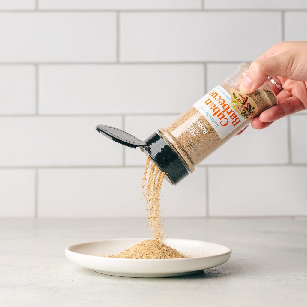 lifestlye shot of Rodelle's cuban barbecue seasoning in the center of the image with cuban roast pork, rice, and beans on two platter. In the background is a plant in a white pot. Utencils also lay on the edges of the image. Table is gray while the background is white